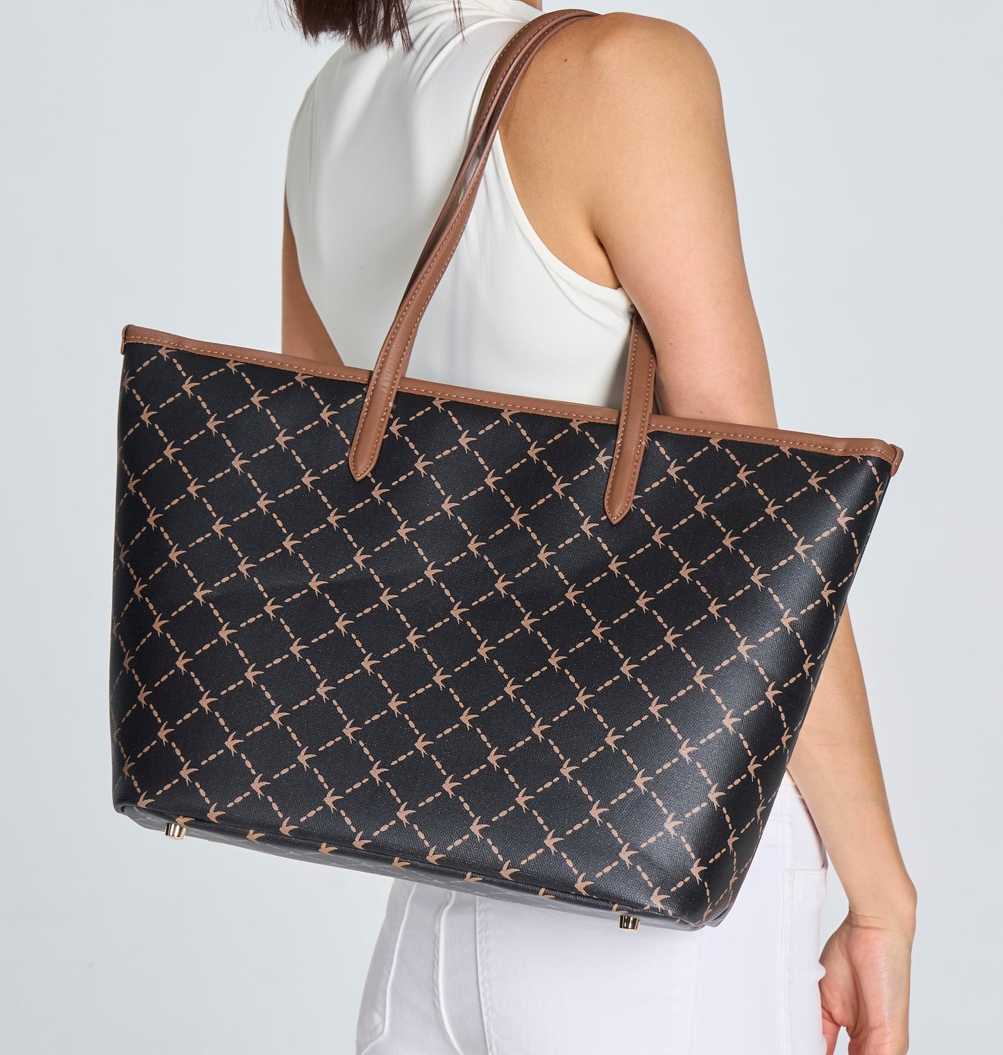 A model carrying a Svala Vita Tote structured vegan work and travel bag with a black base and a repeating brown monogram pattern, featuring brown shoulder straps and trim, shown against a light gray background.