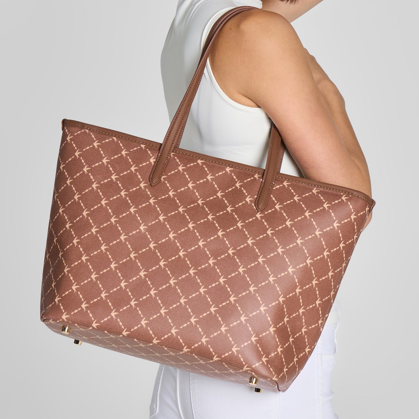 A woman wearing a white tank top carries a large brown vegan work and ravel tote bag with a beige geometric lattice pattern. The shot is cropped from the side/back view against a light gray studio background.