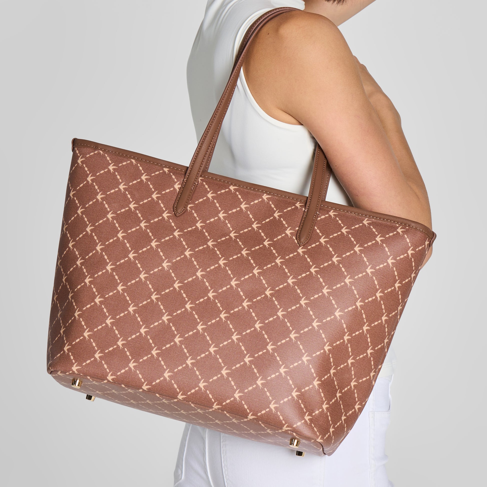 A woman wearing a white tank top carries a large brown vegan work and ravel tote bag with a beige geometric lattice pattern. The shot is cropped from the side/back view against a light gray studio background.