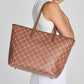 A woman wearing a white tank top carries a large brown vegan work and ravel tote bag with a beige geometric lattice pattern. The shot is cropped from the side/back view against a light gray studio background.